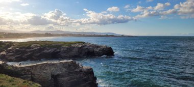 Güneş batarken okyanus dalgalarıyla çerçevelenmiş yeşil tepeler ve kayalar.