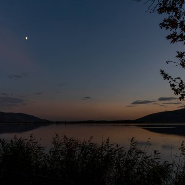 Yunanistan 'ın kuzeyinde sakin bir Orestiada Gölü üzerinde gün batımı önlerinde bataklık çimenleri ve gökte ay