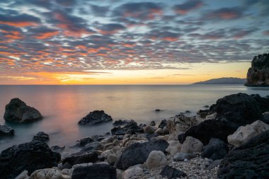Cala Gonone sahilinde renkli bir gün doğumu. Önünde siyah beyaz kayalar ve kayalar var.