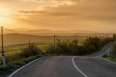 Tuscany 'nin kıvrımlı tepelerinde gün doğumunda elektrik hattı ve sokak tabelası olan kıvrımlı ve tepelik bir ülke otoyolu.