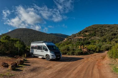 Elba 'nın yeşil tepelerinde mavi gökyüzünün altında toprak bir yolda park etmiş gri bir karavan.