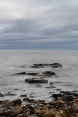 Bulutlu bir gökyüzünün altında dikey deniz manzarası dalgalar ve kayalar ön planda