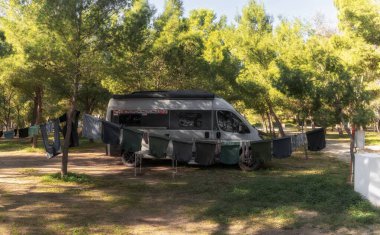 Kamp alanında park etmiş gri bir karavan. Etrafı ağaçlar ve giysilerle çevrili. Ön planda çamaşır ipine asılı.