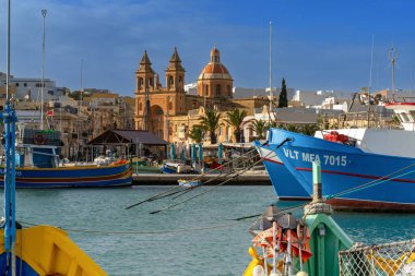 Marsaxlokk, Malta - 22 Aralık 2023: Marsaxlokk şehir merkezi manzaralı kilise ve renkli balıkçı tekneleri ön planda