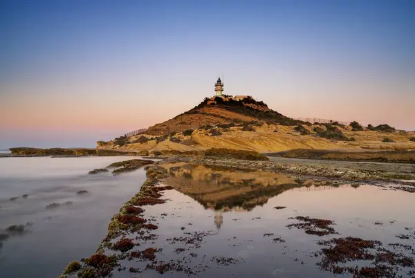 A view of the Cabo de la Huerta lighthouse at sunrise with reflections in tidal pools in the foreground