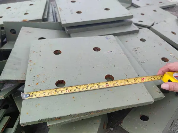Önceden delinmiş deliklerle metal bir plakanın boyutlarını kontrol etmek için bant metresi kullanan eller, inşaat çalışmalarının doğruluğunu vurguluyor.