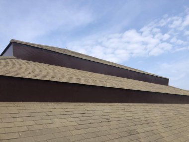 Parlak mavi gökyüzünün altındaki aşınmış asfalt kiremitli çatı, görünür hasar ve çatı yapısında yaşlanma belirtileri gösteriyor