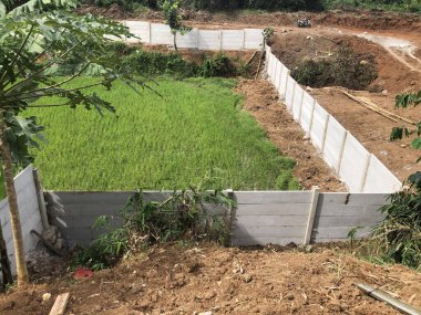 Etrafı tropik yeşilliklerle çevrili açık bir alanı çevreleyen uzun beton çitler.