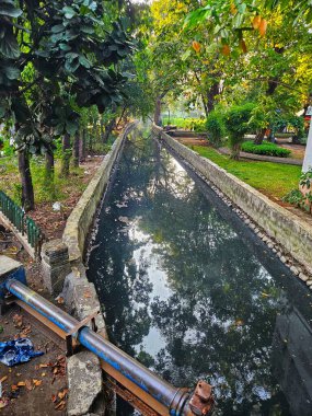 Sakin, insan yapımı bir su kanalı yemyeşil bir parkın içinden akıyor, sakin gökyüzü ve yemyeşil ağaçlar suya yansıyor.