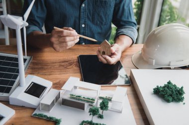 Mimar tasarımcı, ev tasarımıyla doğal enerji kavramını birleştirmeyi düşünürken küçük ahşap bir ev sembolü tutuyor. Modern ev ve rüzgar türbini modeli, masa başında güneş panelleri..