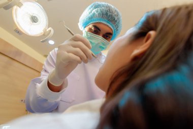 Maskeli ve eldivenli dişçi. Diş sağlığı kliniğinde hastanın dişlerinde çürük var mı diye kontrol ediyor..