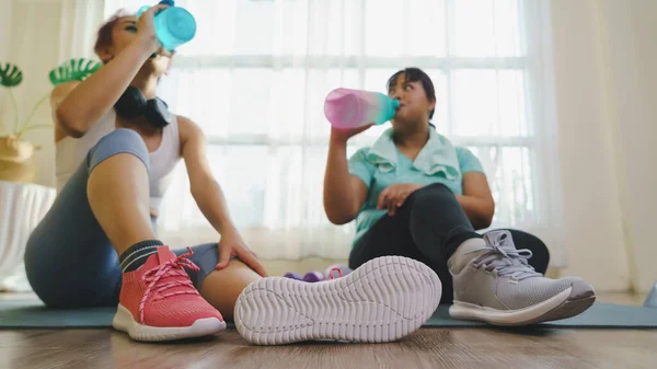 Thirst while exercising concept. Two Asian women\'s body size is different in sportswear, drinking water while resting and sitting on yoga mat after fitness exercise at home together.