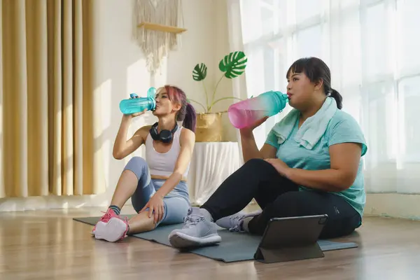 Thirst while exercising concept. Two Asian women\'s body size is different in sportswear, drinking water while resting and sitting on yoga mat after fitness exercise at home together.