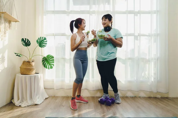 Enjoy fitness and Healthy food concept. Two Asian women body size different in sportswear standing while smiling enjoy fitness and happy in eating healthy food after exercising at home together.