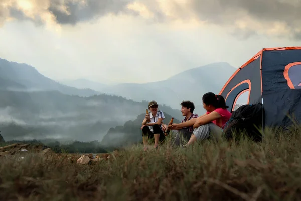 Asian young people groups sit enjoy with beer and drinking near a camping tent with mountain views of natural rainforest in foggy weather. Enjoying nature with soft light rain in background