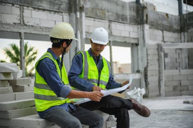 İnşaat mühendisleri ustabaşı veya inşaatçıyla planları açarken ve inşaat alanındaki tuğla yığınlarının üzerinde otururken tartışırlar..
