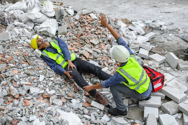 Accident in construction workplace, Knee accident from slip or stumble fall on the concrete scrap at construction site. Foreman helping injured colleague with first aid bag and wave hand to team help