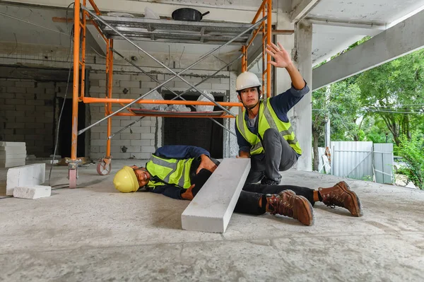 Accident in the construction workplace, concrete block brick falls on an unsuspecting builder\'s leg, Foreman to helping injured colleague or builder and wave hand to safety officer for help.