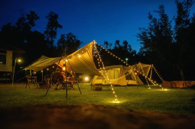 Açık hava kamp çadırı, çimen bahçesinde muşamba ya da uçan çarşaf, karanlık mavi gökyüzü alacakaranlık vakti altında sıcak gece ışığı, tatilde aile pikniği, tatilde dinlenme, aile turistlerinin kamp yapma manzarası..