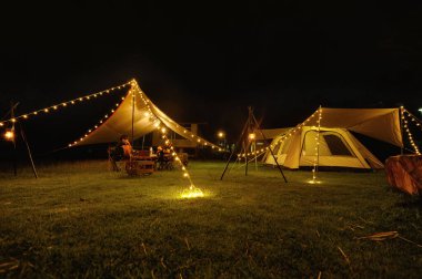 Açık hava kamp çadırı, çimen avlusunda muşamba ya da sinek örtüsü ve karanlık gece gökyüzünün altında sıcak gece ışığı, tatilde aile pikniği, tatilde dinlenme, aile turistlerinin kamp manzarası..