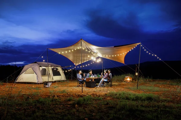 Outdoor camping tent in the forest park, party dinner with friends under tarp or flysheet and warm lighting at night near natural under a blue twilight sky. Overview of travel camping with friends.