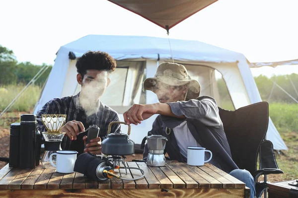 Asian young man two persons sitting enjoy making drip coffee drink near a camping tent, coffee drink relaxing on the morning summer camping vacation.