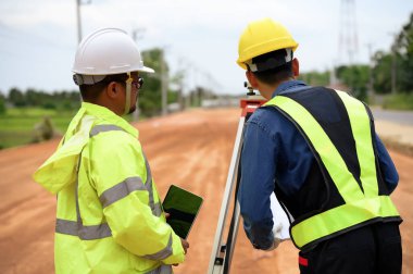 Asyalı arazi mühendisi iki kişi kesim ve dolgu seviyesini ölçmek için Teleskop ekipmanıyla toprağı kontrol ediyor, karayolu inşaat sahasının zeminini düzleştirmeye başladı.