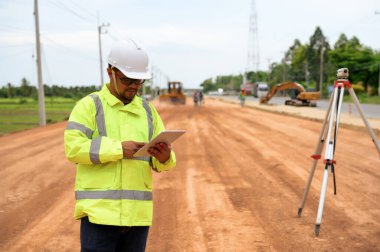 Asyalı arazi mühendisi teleskop seviyelerine yakın duruyor ve dijital çizim seviyesinin detaylarını kontrol etmek için teknoloji tableti kullandı. Otoyol inşaatındaki kesim ve dolgu seviyesini ölçmek için..