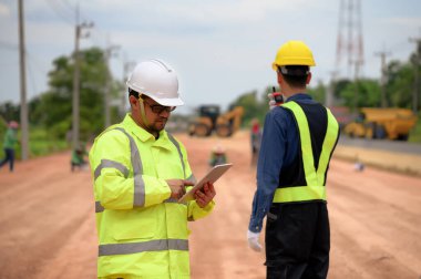 Asyalı mühendisler iş yerinde ayrıntıları kontrol etmek için tablet, inşaat ekibiyle karayolu yapım projelerinin çalışma saatlerini kontrol etmek ve arka planda makineler inşa etmek için tablet kullanırlar.