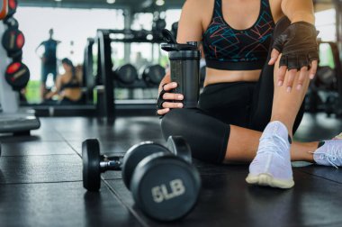 Spor salonundaki dambıllarla spor yaptıktan sonra dinlenen ve protein içeceği şişesini sağlıklı yaşam tarzında tutan kadın. Kadın konseptinin kas geliştirme egzersizi.