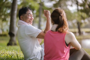 Asyalı Yaşlı Baba ve Kızı sıcak akşam ışığıyla yeşil bir parkta dinleniyorlar. Temiz havayı solumak ve güzel manzaranın tadını çıkarmak. Sağlık ve aile bağları.