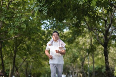 Asyalı yaşlı mutlu adam yeşil ağaçlarla dolu bir parkta tek başına koşuyor..