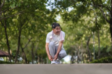 Yaşlı bir Asyalı adam nazik bir yüzle ayakkabı bağcıklarını bağlıyor ve parkta nazik bir gülümseme ağaçlarla dolu. Parka bakıyor ve gülümsüyor Mutlu ve canlı hissederken