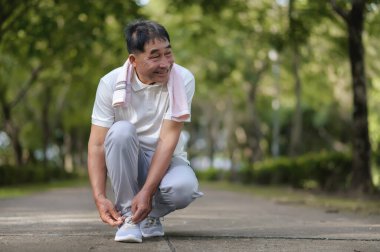 Yaşlı bir Asyalı adam nazik bir yüzle ayakkabı bağcıklarını bağlıyor ve parkta nazik bir gülümseme ağaçlarla dolu. Parka bakıyor ve gülümsüyor Mutlu ve canlı hissederken