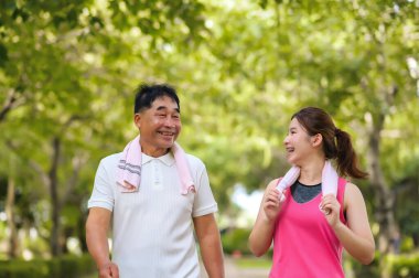 Asyalı Yaşlı Baba ve Kızı yeşil bir parkta rahatça yürüyor, temiz havayı soluyor ve güzel manzaranın tadını çıkarıyorlar. Sağlık ve aile bağları.