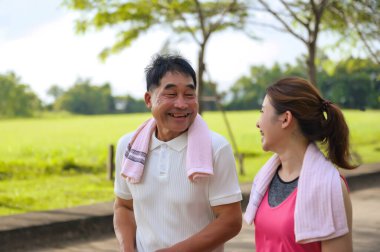 Asyalı Yaşlı Baba ve Kızı yeşil bir parkta rahatça yürüyor, temiz havayı soluyor ve güzel manzaranın tadını çıkarıyorlar. Sağlık ve aile bağları.