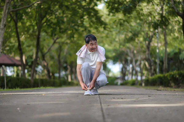 An Elderly Asian Man Is Tying Shoelaces With A Kind Face And A Gentle Smile In The Park Is Full Of Trees. Looks Around The Park and Smiles While Feels Happy And Alive