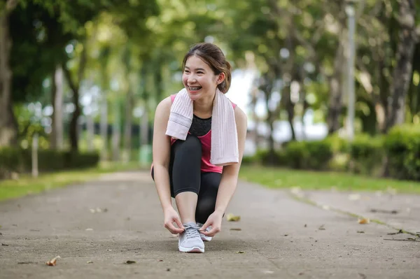 An Asian Woman Is Tying Shoelaces With A Kind Face And A Gentle Smile In The Park Is Full Of Trees. Looks Around The Park and Smiles While Feels Happy And Alive