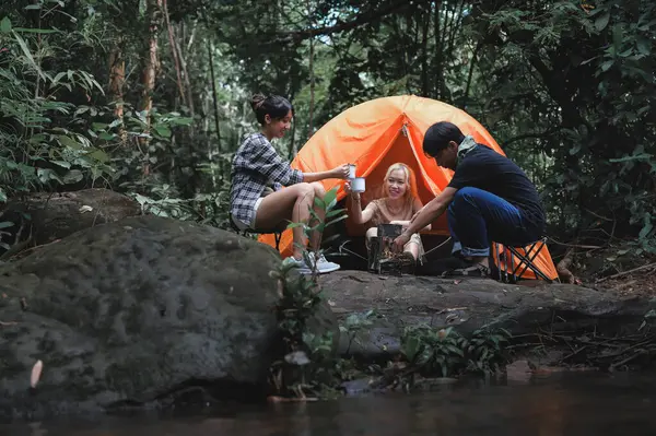 Young People Trekking Group Sitting Relaxed in the Rainforest Near a Natural Stream and Boil Water with a Camp Stove. Campsite Drinking Coffee and Water. Prepare Camping Meals for Groups on Trip.
