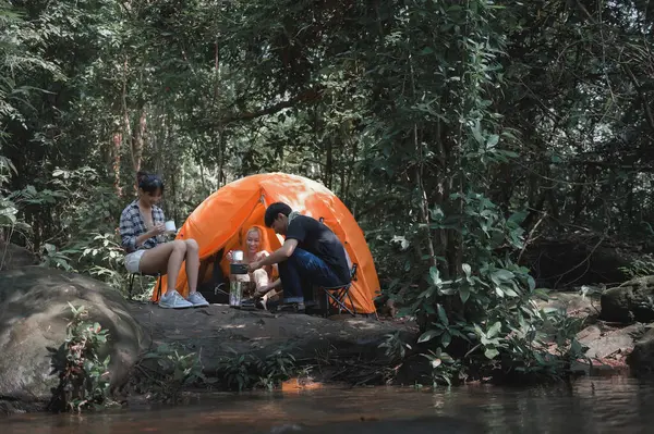 Young People Trekking Group Sitting Relaxed in the Rainforest Near a Natural Stream and Boil Water with a Camp Stove. Campsite Drinking Coffee and Water. Prepare Camping Meals for Groups on Trip.