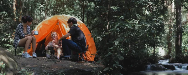 Young People Trekking Group Sitting Relaxed in the Rainforest Near a Natural Stream and Boil Water with a Camp Stove. Campsite Drinking Coffee and Water. Banner Size with Copy Space.