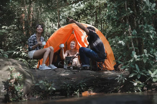 Young People Trekking Group Sitting Relaxed in the Rainforest Near a Natural Stream. Looking at Natural Views in the Forest while Campsite Drinking Coffee and Water. Prepare Camping Meals for Groups.