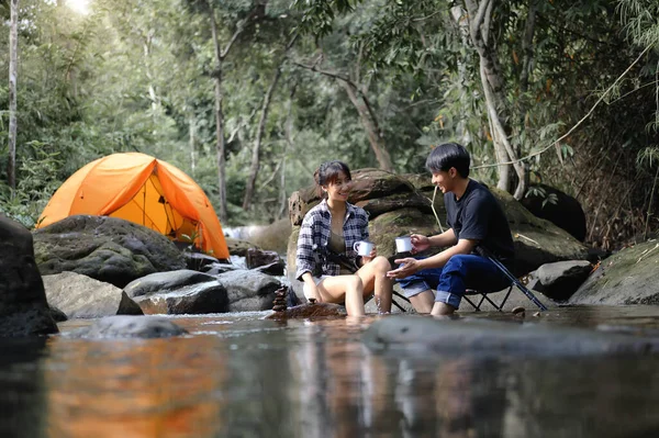 Asian Teenager sitting on a camping chair in the natural stream with a camping tent in the background, Enjoying sitting relaxed and drinking water in the atmosphere of the surrounding nature.