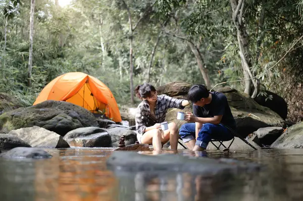 Asian Teenager sitting on a camping chair in the natural stream with a camping tent in the background, Enjoying sitting relaxed and drinking water in the atmosphere of the surrounding nature.
