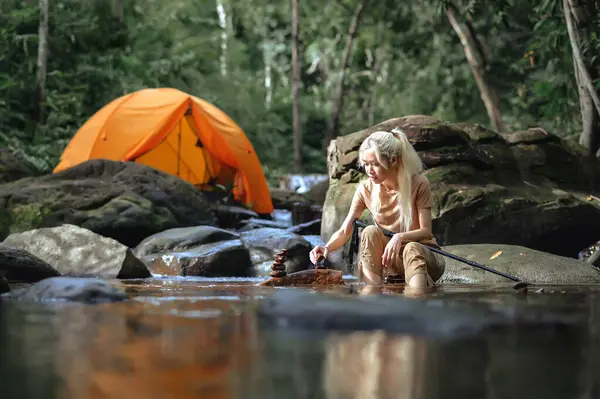 Asian woman sitting on a camping chair in the natural stream with a Camping tent in the background, Enjoying building balanced natural stone arrangements. Healthy lifestyle by nature therapy.