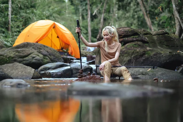 Asian woman sitting on a camping chair in the natural stream with a Camping tent in the background, Enjoying building balanced natural stone arrangements. Healthy lifestyle by nature therapy.