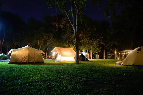 Outdoor Glamping tent at night on grass courtyard and warm night light under dark blue sky twilight time, Group of family vacation camp on holiday relax, Overview of camping of family tourist.