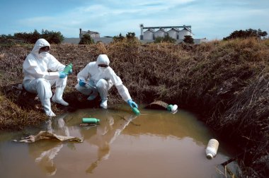 Bilim adamları ya da biyologlardan oluşan bir ekip, doğal su kaynağındaki kimyasal dolu çöp şişelerini incelemek ve not etmek için koruyucu giysiler giyer. Su kirliliği kavramı.