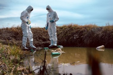 Bilim adamları ya da biyologlardan oluşan bir ekip, kimyasal dolu çöp şişeleri olan doğal bir su kaynağından su örnekleri toplamak için koruyucu giysiler giyer. Su kirliliği kavramı.