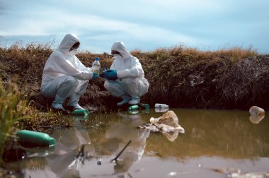Bilim adamları ya da biyologlardan oluşan bir ekip, kimyasal dolu çöp şişeleri olan doğal bir su kaynağından su örnekleri toplamak için koruyucu giysiler giyer. Su kirliliği kavramı.